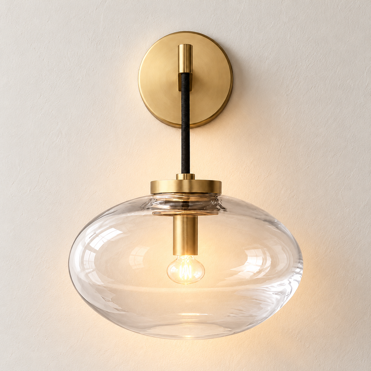 Brass wall sconce with a clear flattened glass shade, illuminated on ivory plaster.