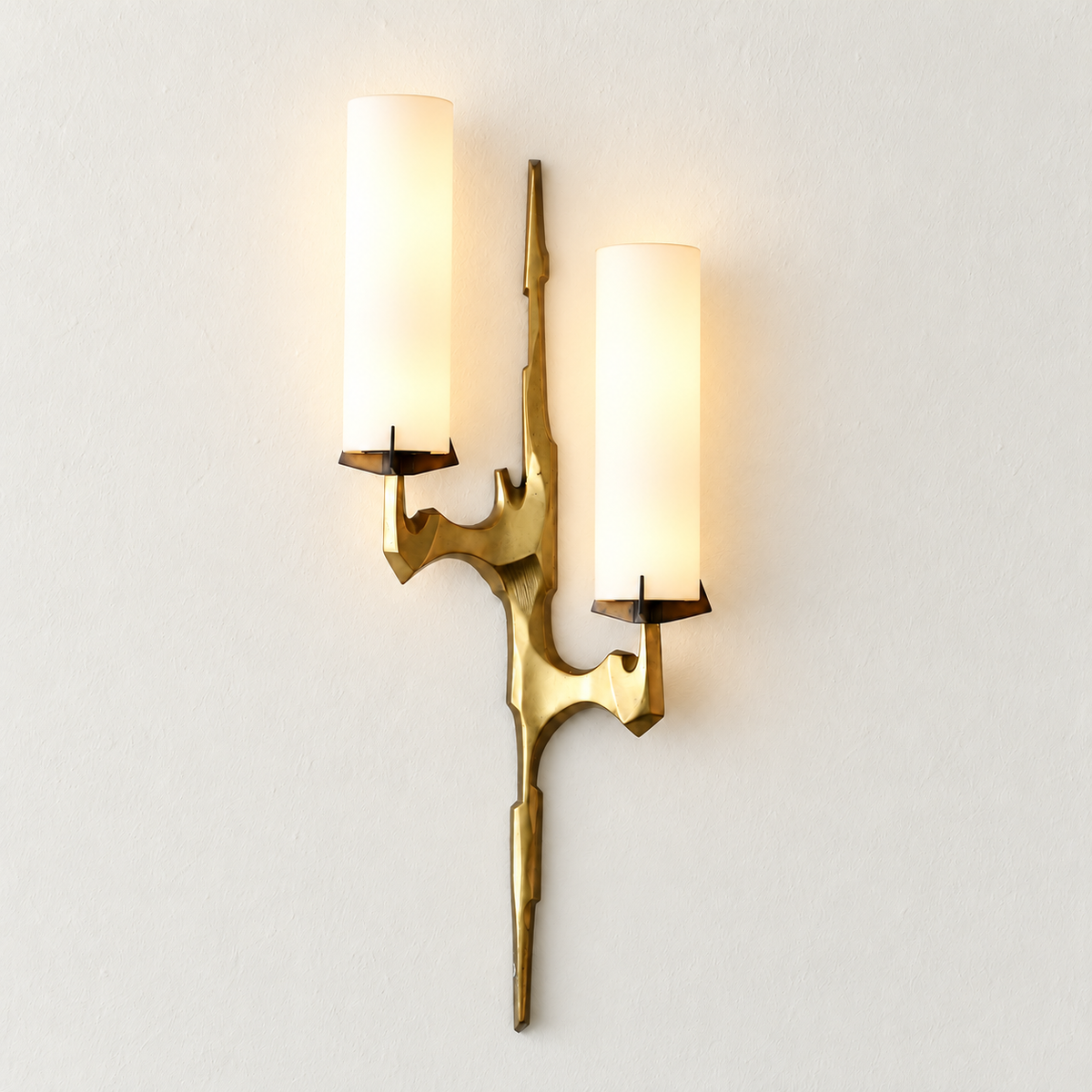 Sculptural brass sconce with two staggered opal glass shades illuminated against the same warm ivory backdrop.
