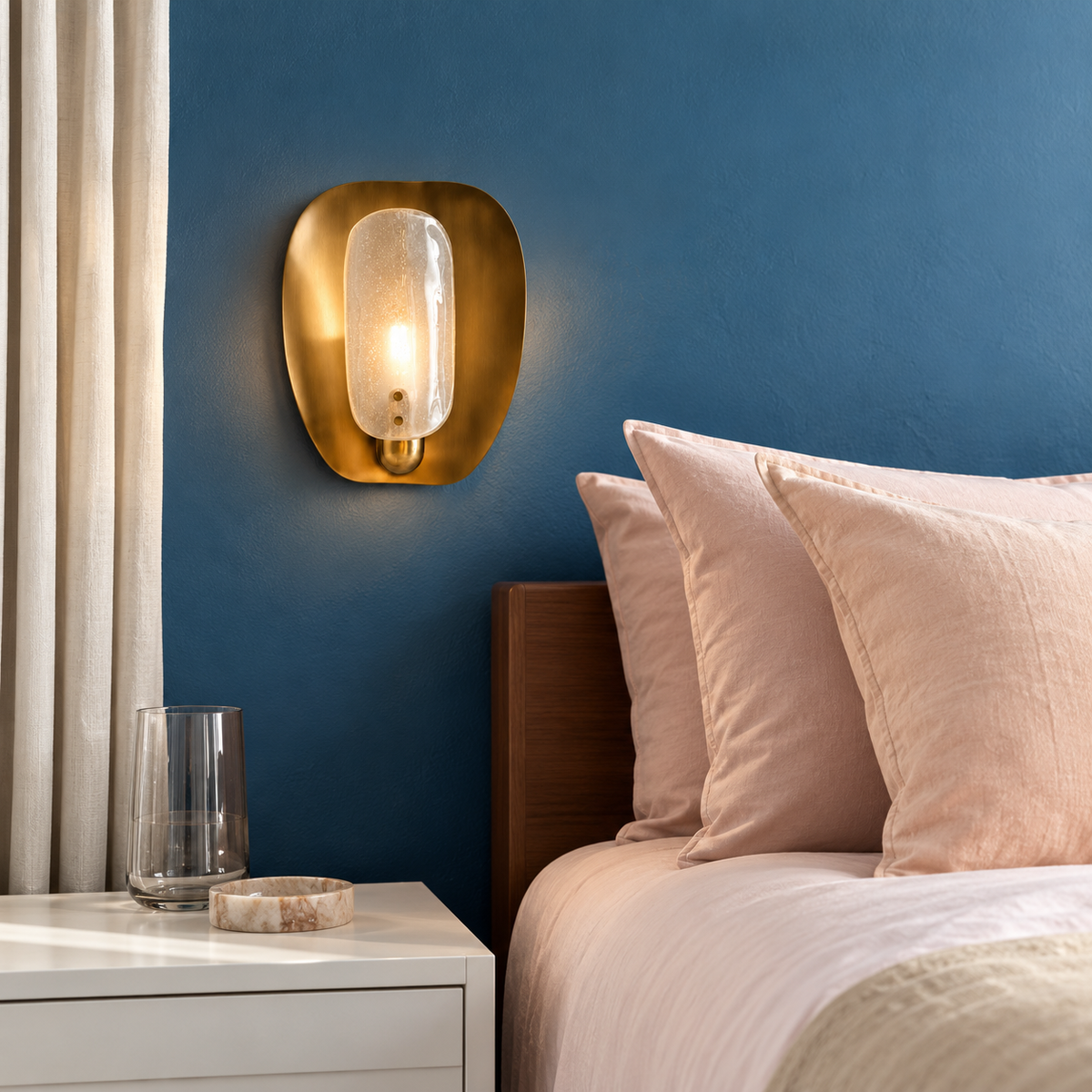 Brass and bubbled glass wall sconce beside blush bedding and an ivory curtain in a blue bedroom.