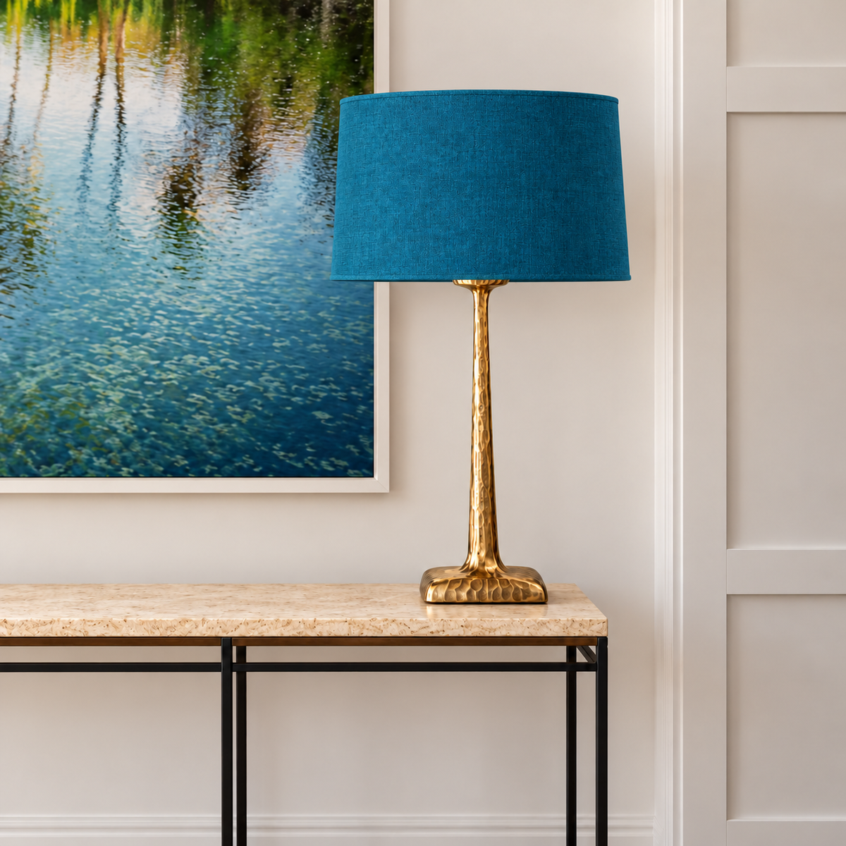Hammered brass table lamp with a petrol blue tapered fabric shade on a stone console beneath framed artwork.