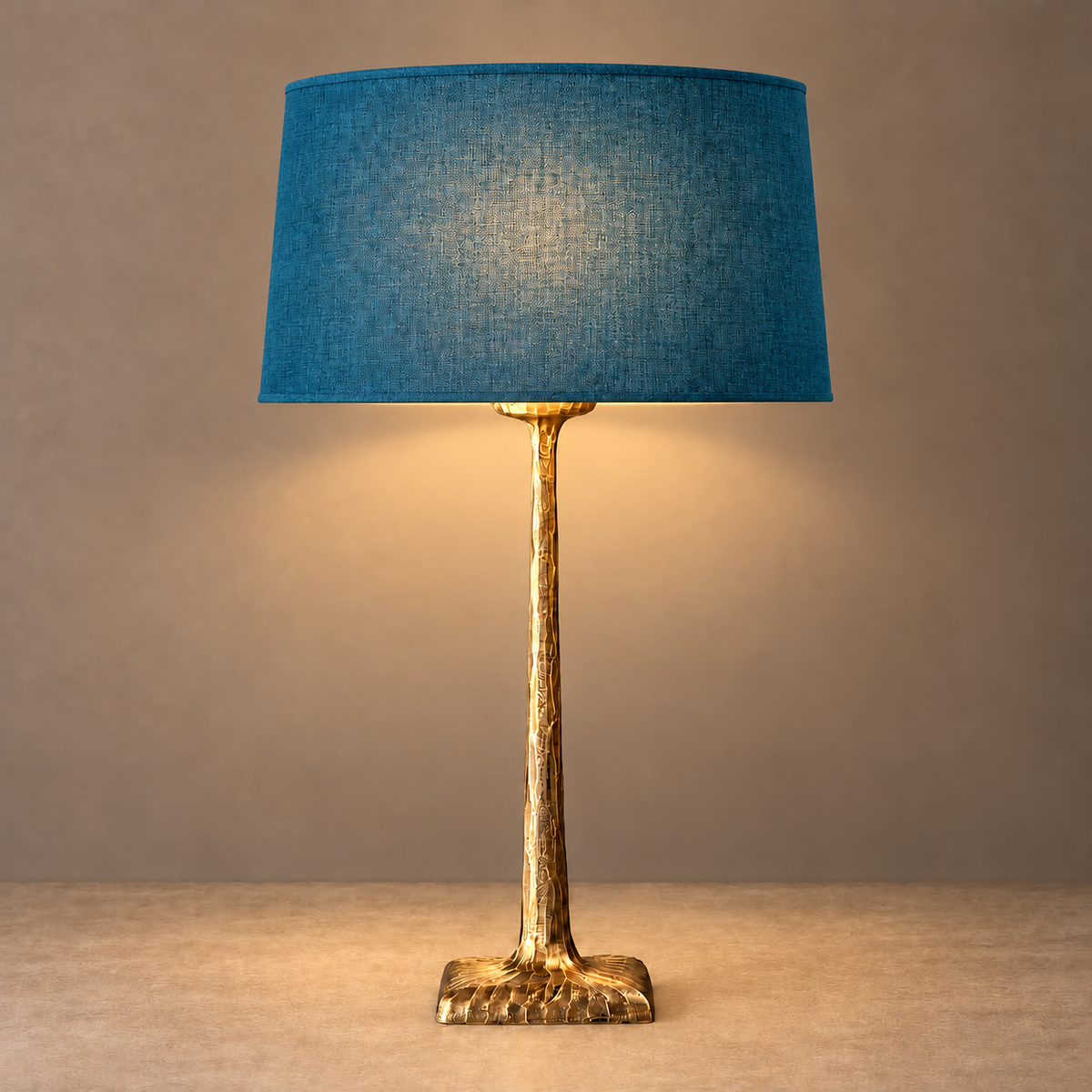 Hammered brass table lamp illuminated beneath its petrol blue tapered fabric shade.