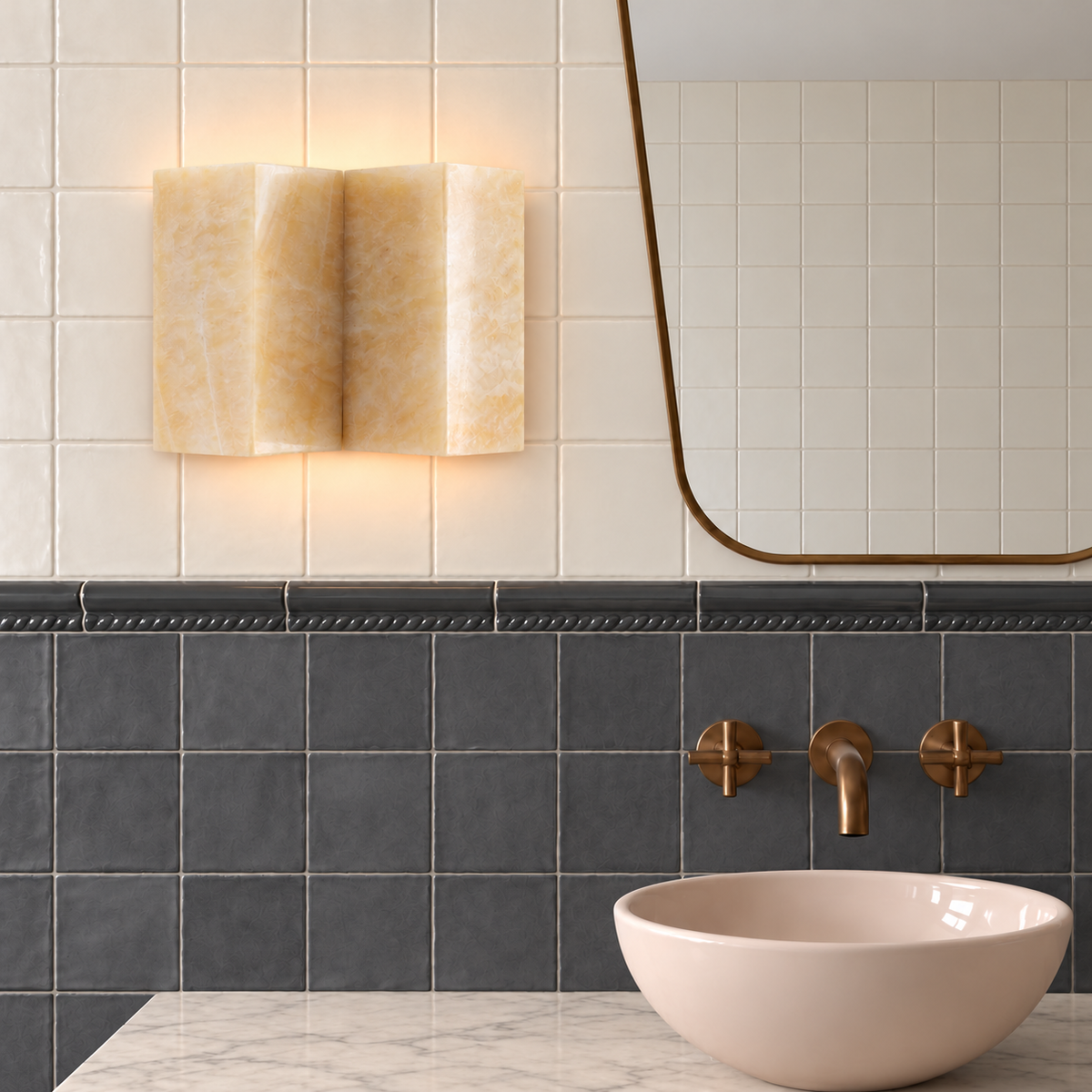 Wide honey onyx wall sconce beside a brass mirror in an ivory and charcoal tiled bathroom.