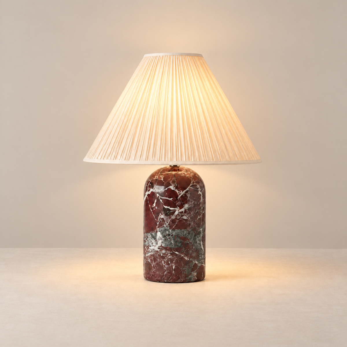Red marble table lamp with its cream pleated linen shade illuminated.