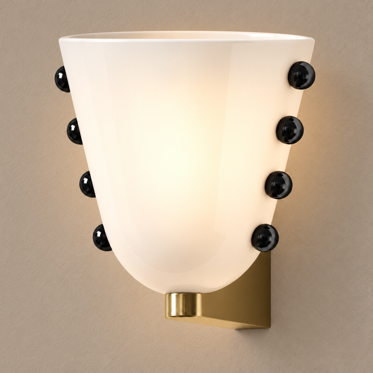 White glass wall sconce with black studs and gold brass mount, illuminated on a stone-beige wall