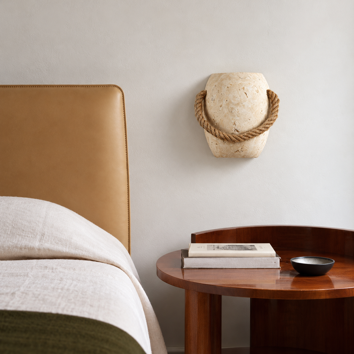 Travertine and rope sconce above a wooden bedside table beside a tan leather headboard.