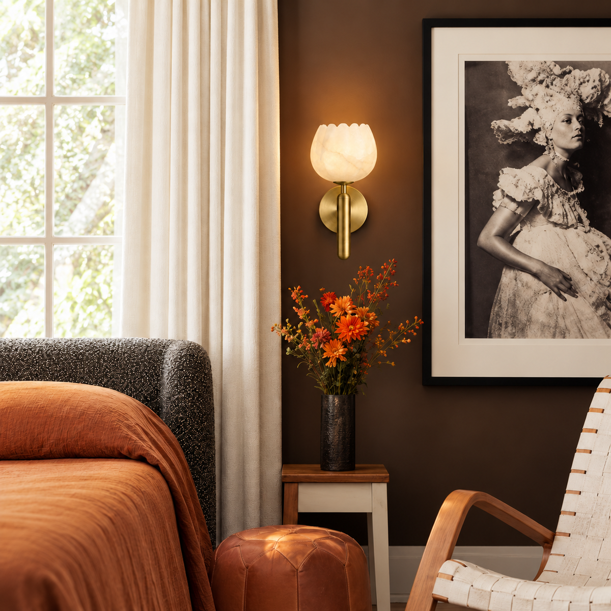 Alabaster tulip and brass wall sconce beside ivory curtains and rust bedding in a brown bedroom.