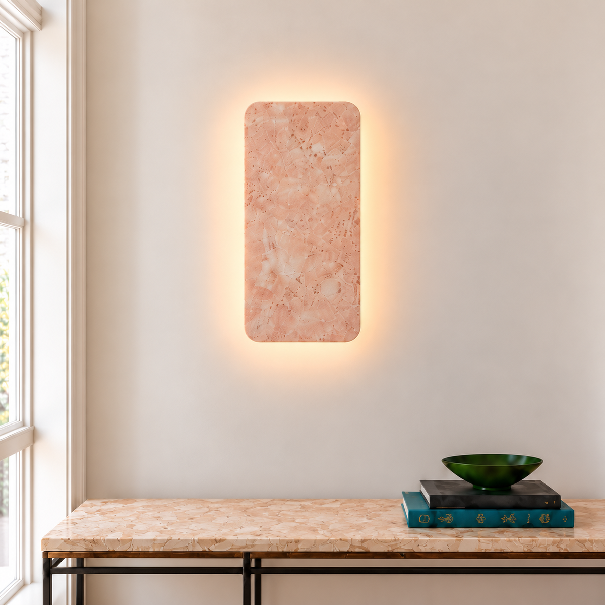 Illuminated rectangular pink marble wall sconce above a marble console with books and a green bowl.