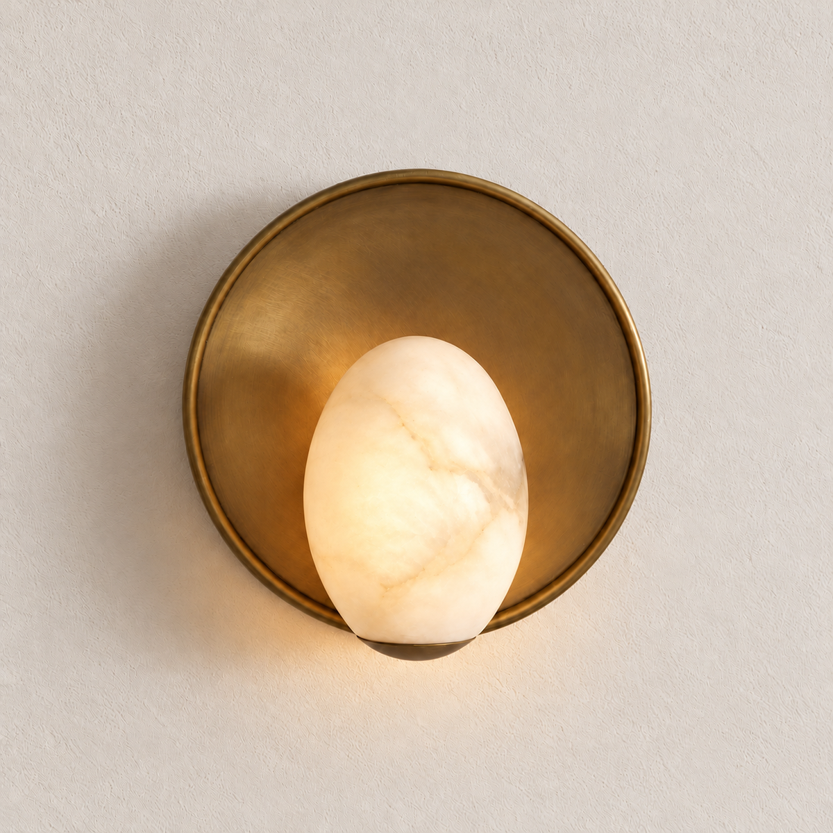 Alabaster egg wall sconce glowing warmly in front of its round brass backplate, shown on warm ivory.