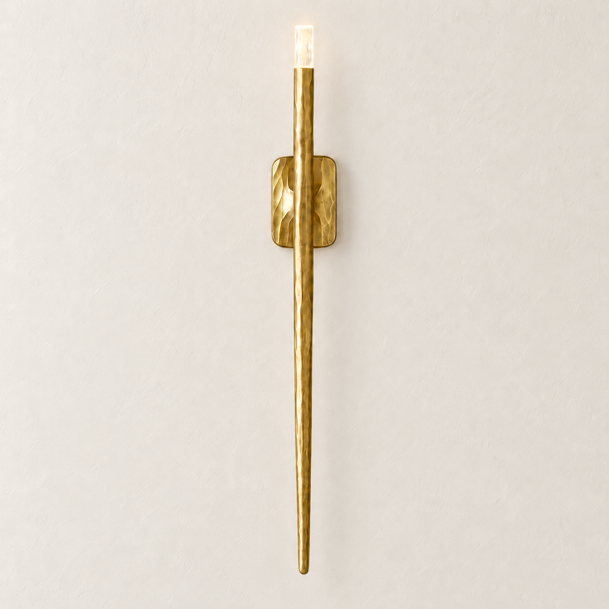Front view of the long textured brass wall sconce, illuminated on ivory plaster.