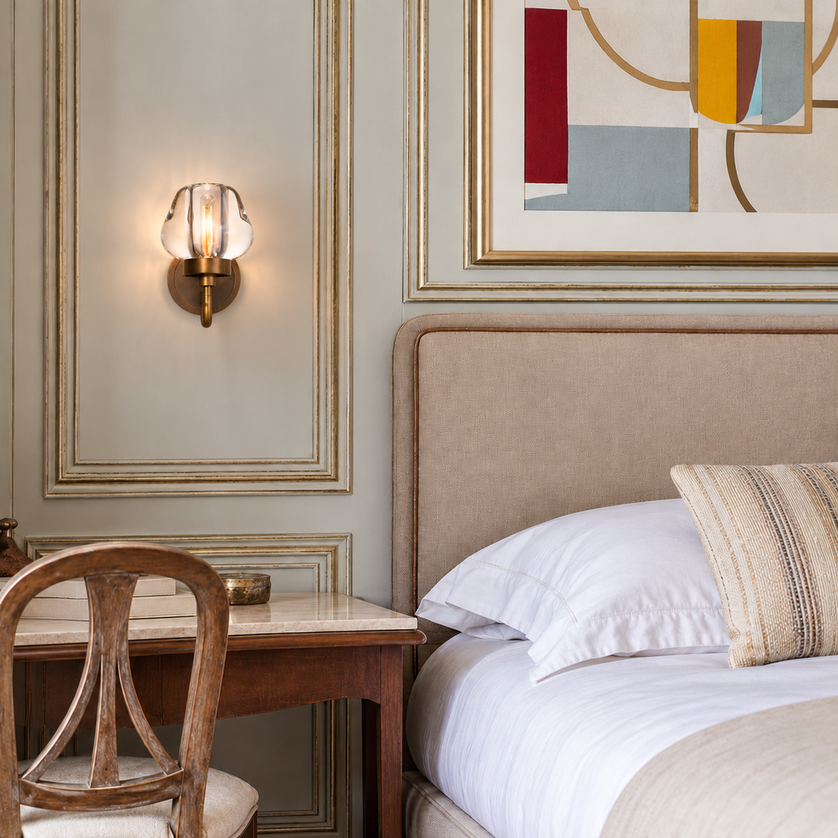 Small brass and crystal wall sconce beside a beige bed in a paneled bedroom.
