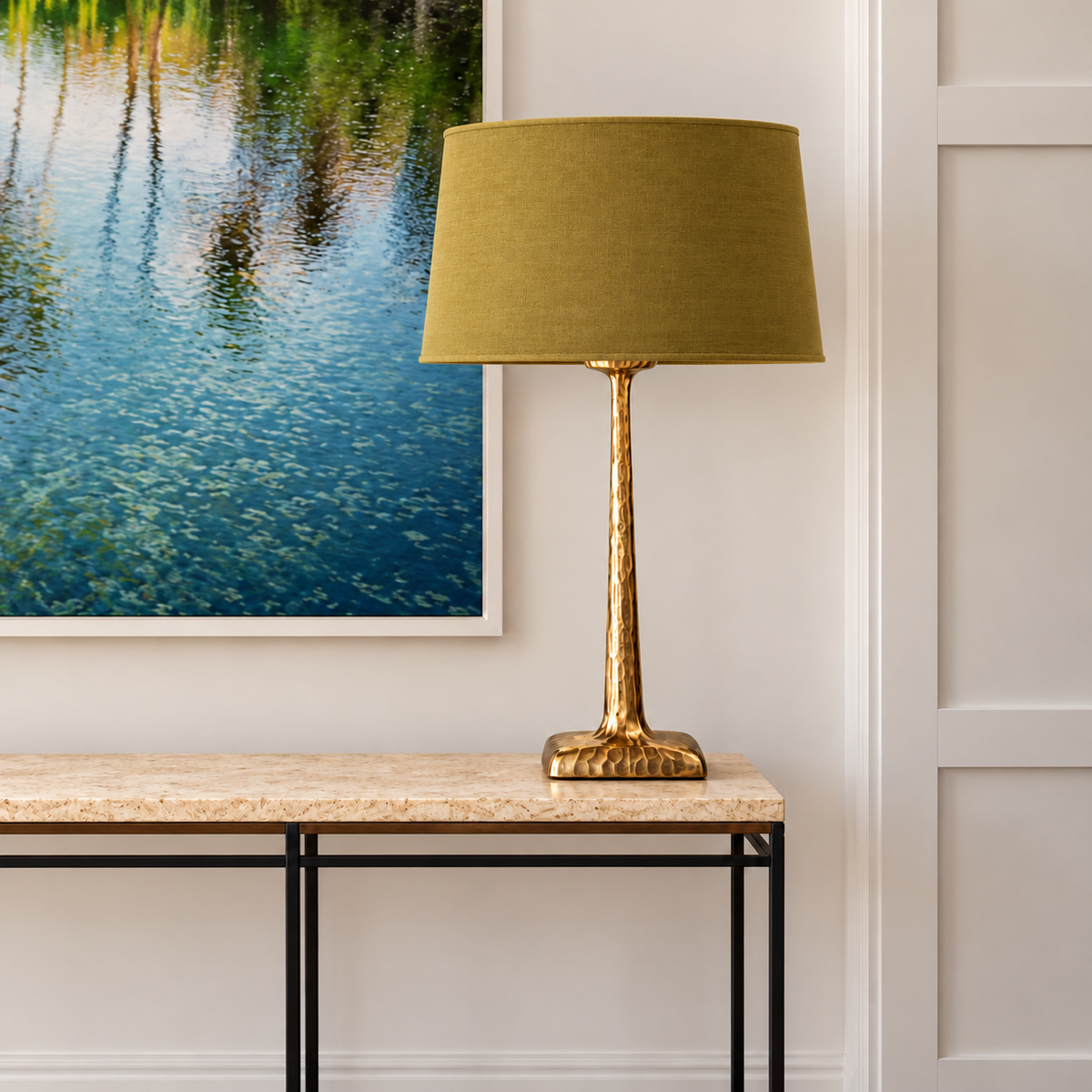 Hammered brass table lamp with an olive tapered fabric shade on a stone console beneath framed artwork.